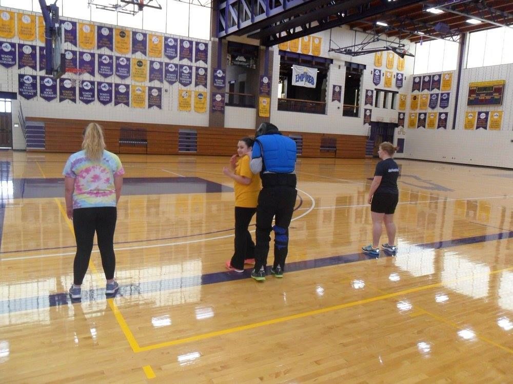 People in a gymnasium. Two people are speaking, while two others stand nearby. Purple and gold banners hang on the wall.