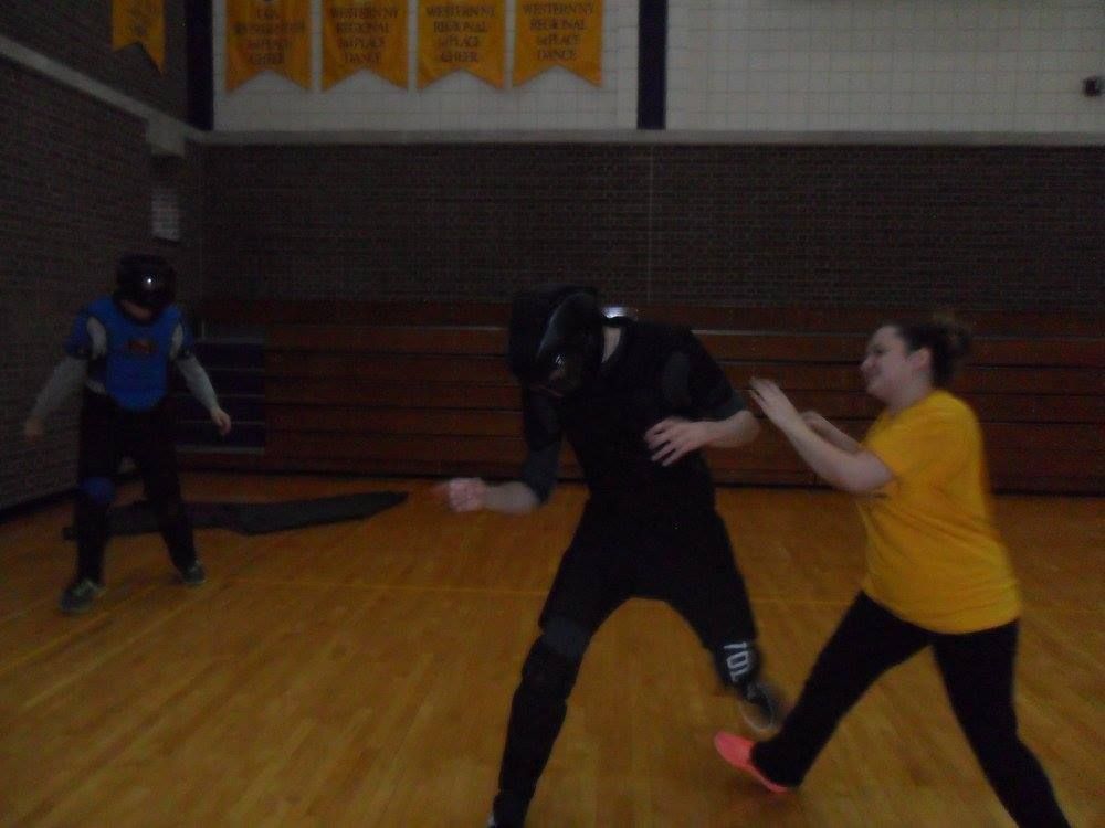 A woman in a yellow shirt kicks a person in black fencing gear during a fencing practice. A third person in blue protective gear is in the background.