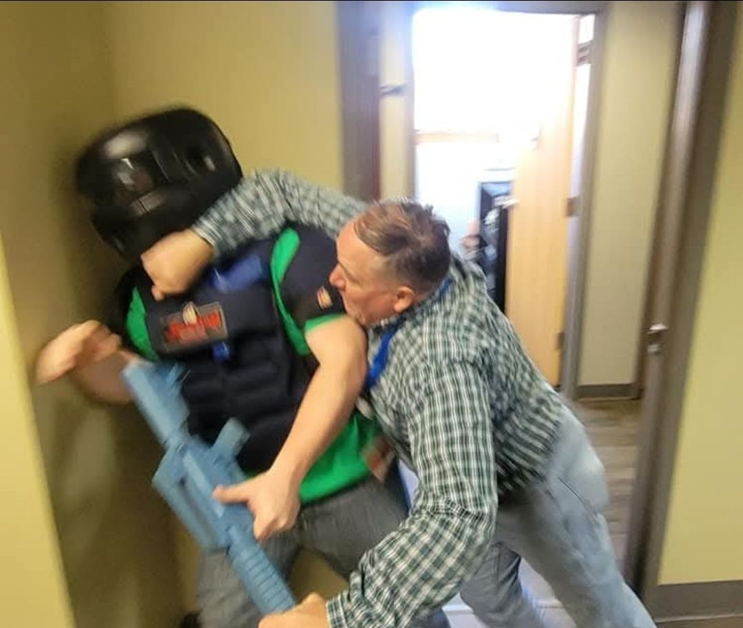 A person in tactical gear is pushed against a doorway by another person in a building hallway.