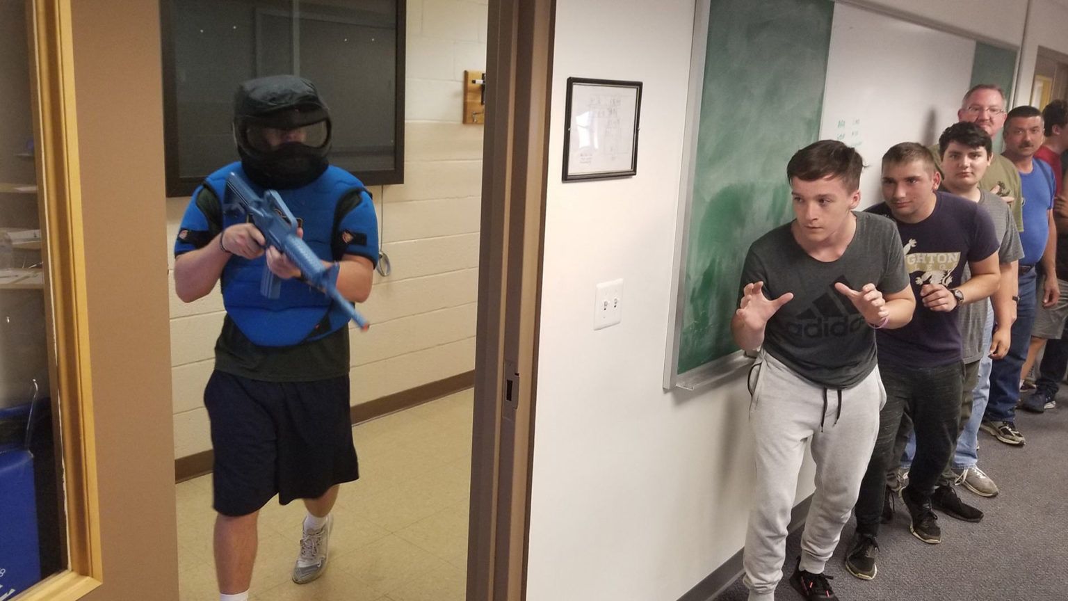 A person in tactical gear holding a blue rifle enters a hallway where people are lined up against the wall, showing expressions of fear.