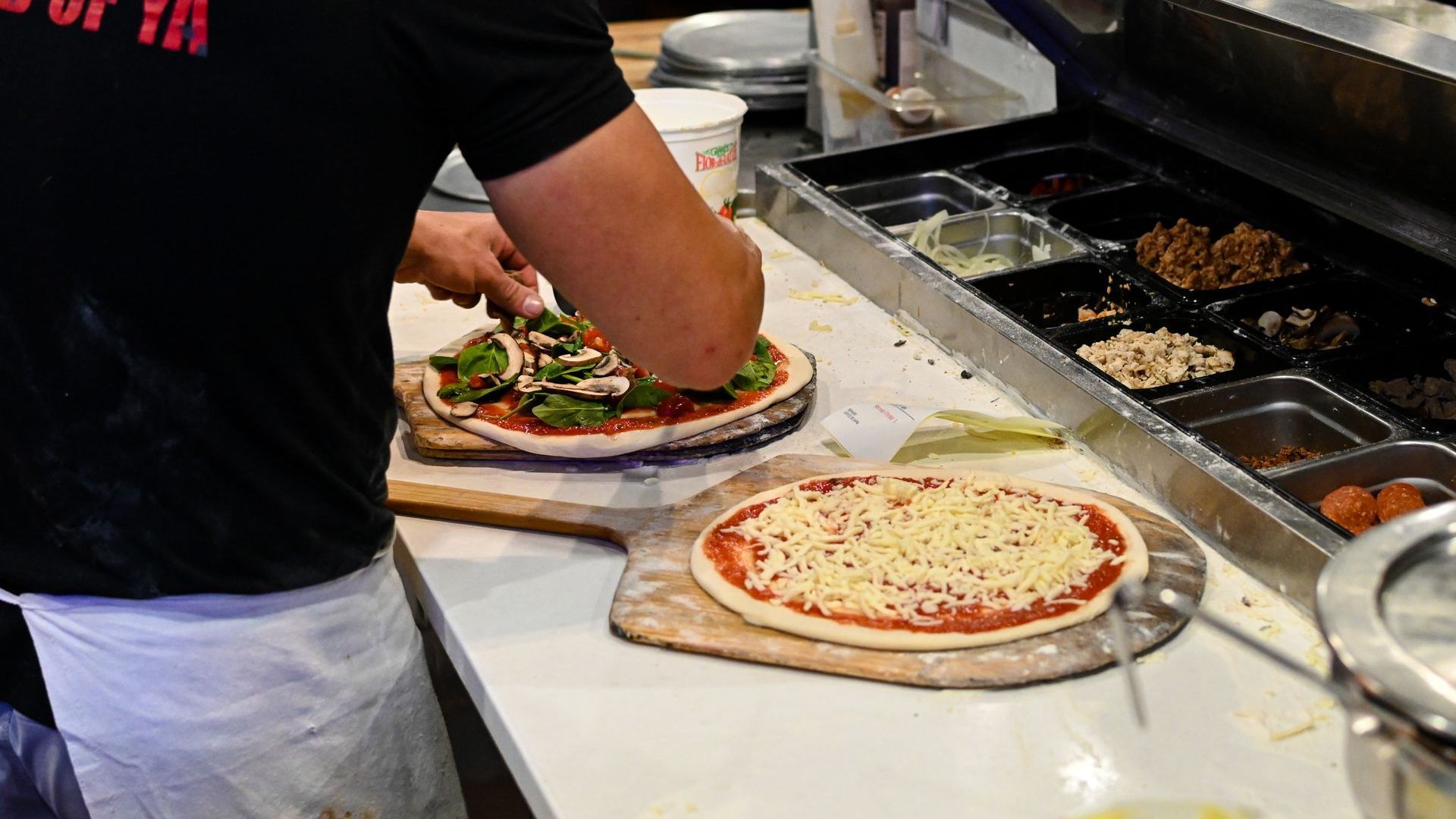 Person topping two pizzas with ingredients at a counter in a restaurant.