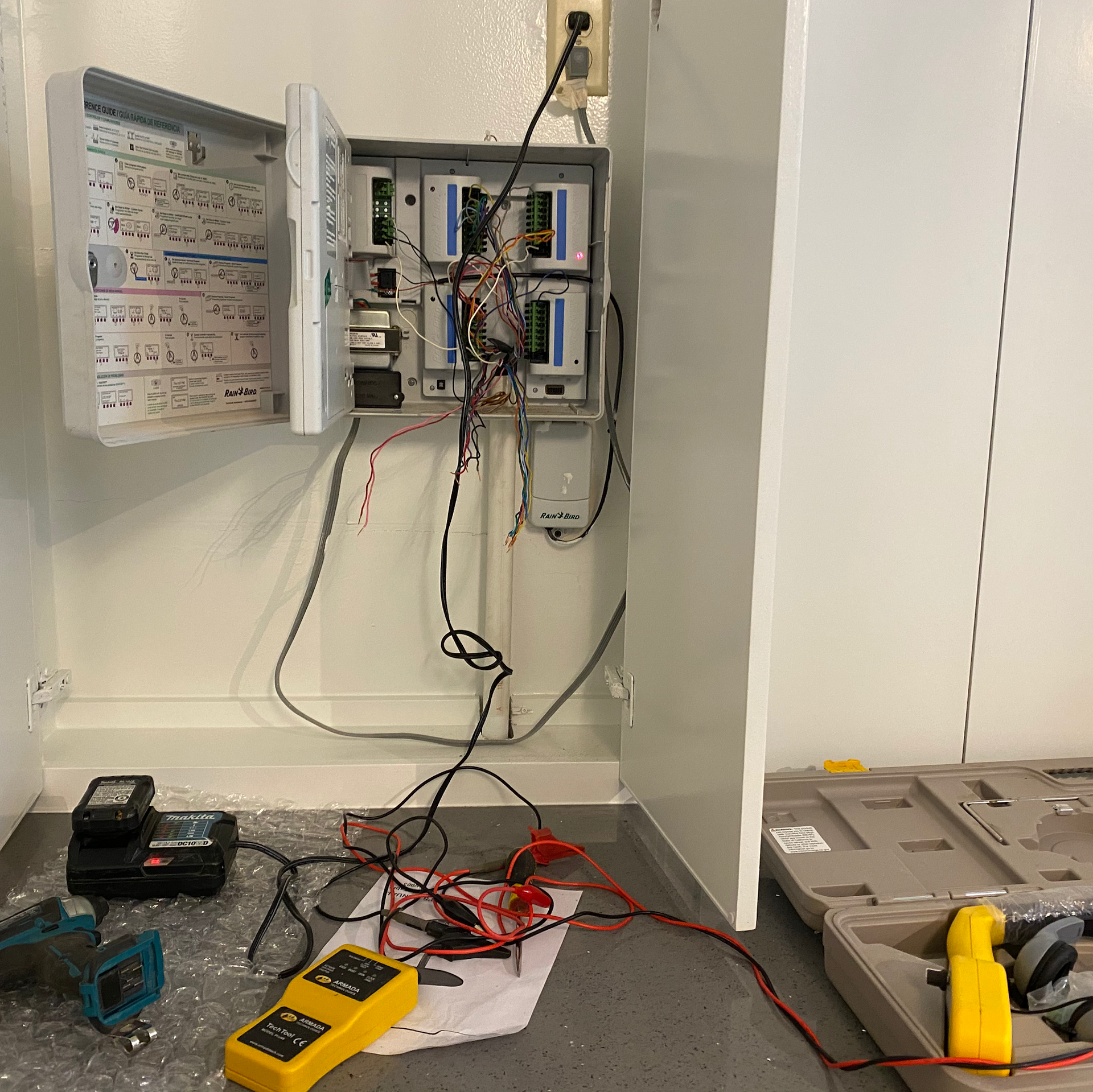 Electrical panel with open door, wires, and testing equipment inside a white-walled room.