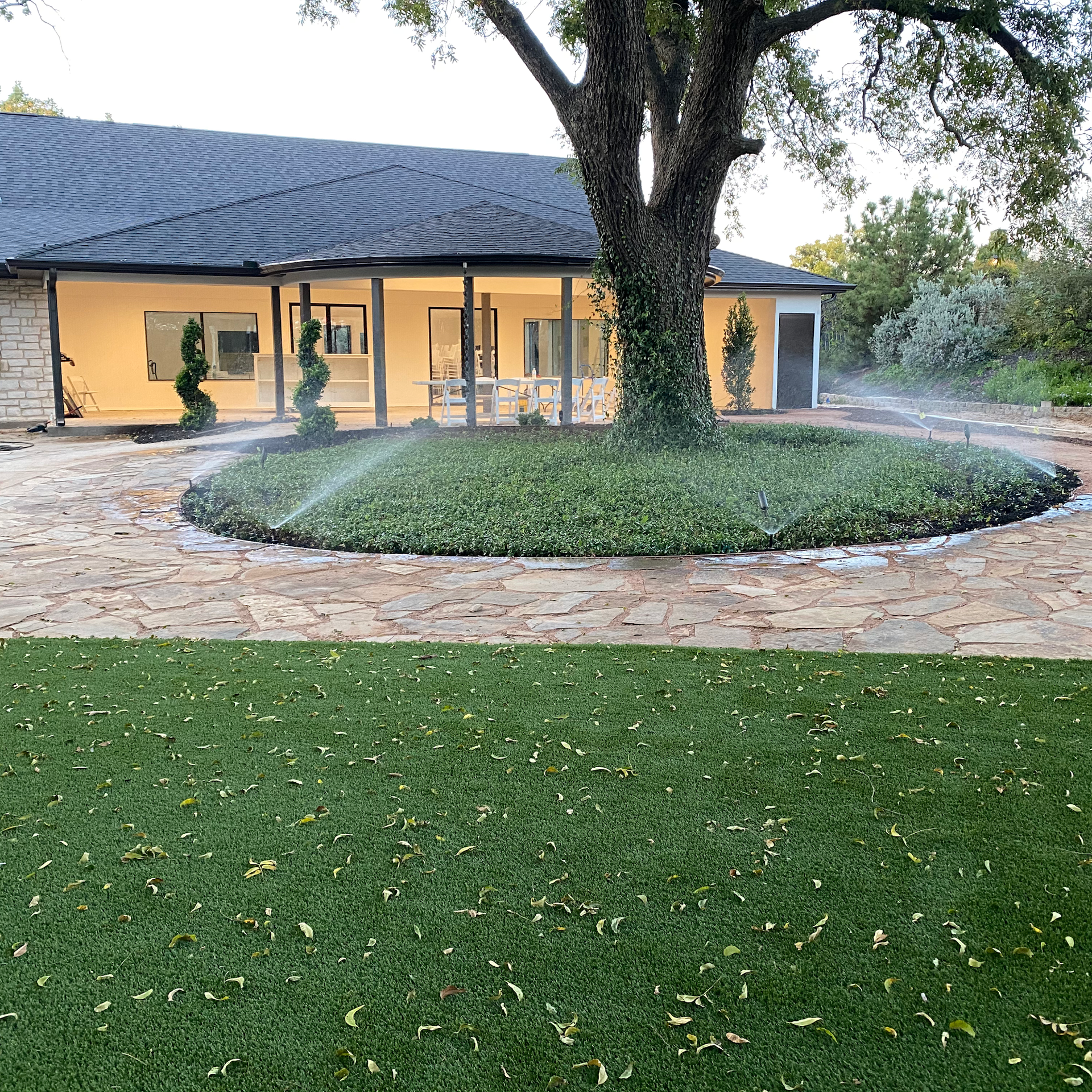 House with stone path, green lawn, and circular bed of greenery around a tree; sprinkler system active.