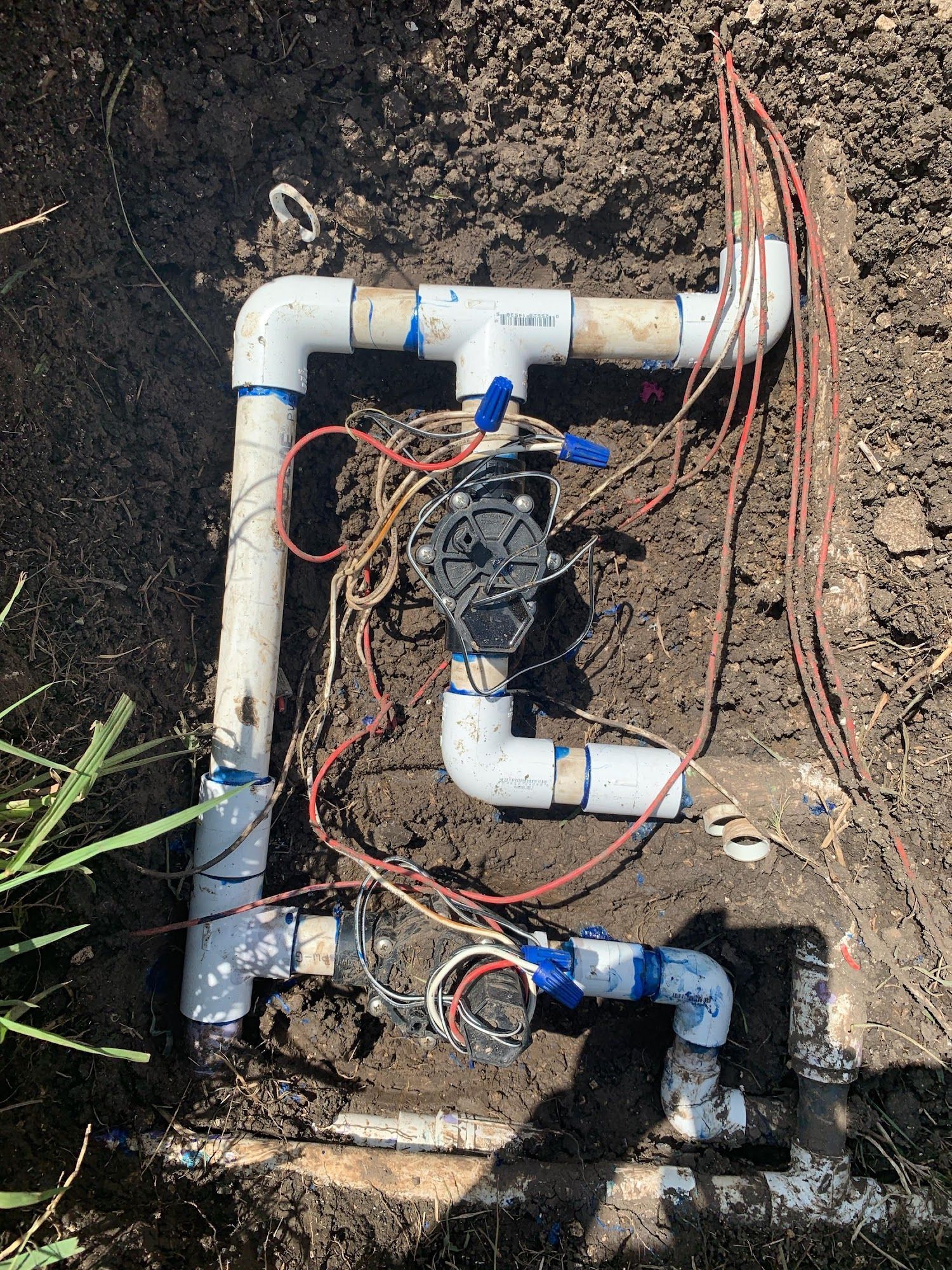 Irrigation system components buried in dirt, including white PVC pipes, valves, and wiring.