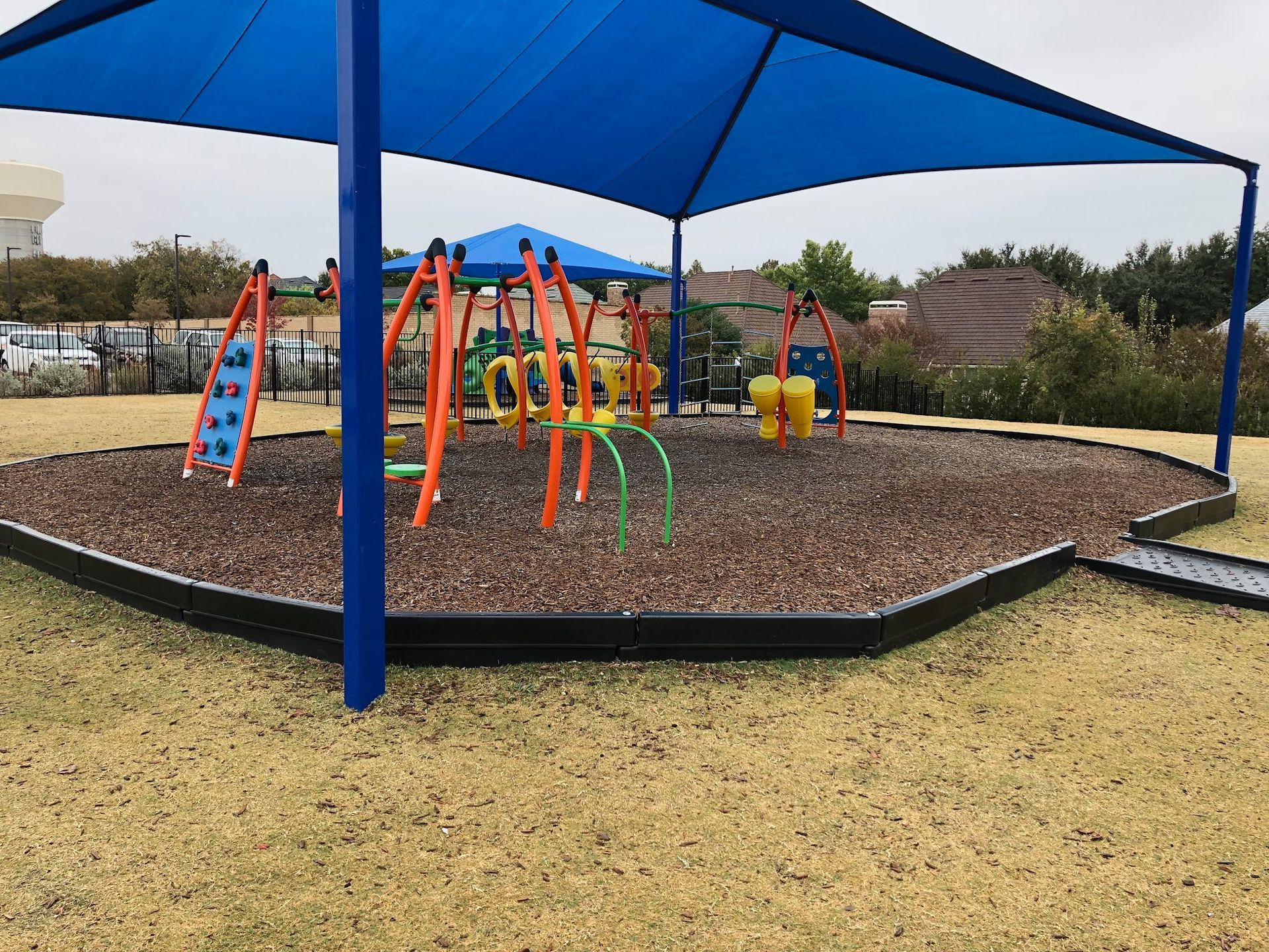 Playground with blue shade canopy and colorful equipment on a mulch base, set on grass.