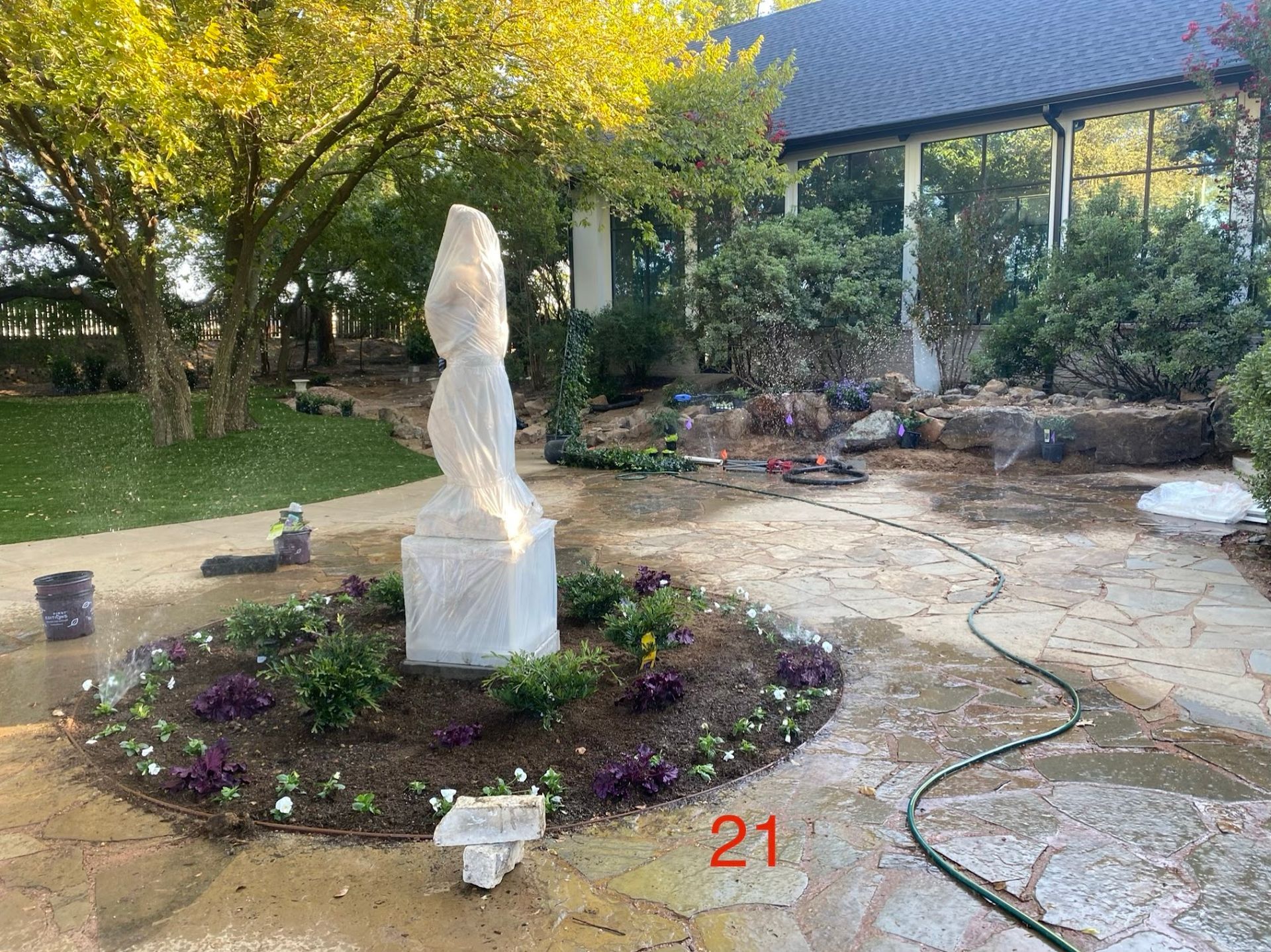 A white statue on a flowerbed in a garden. A hose lies on a stone path.