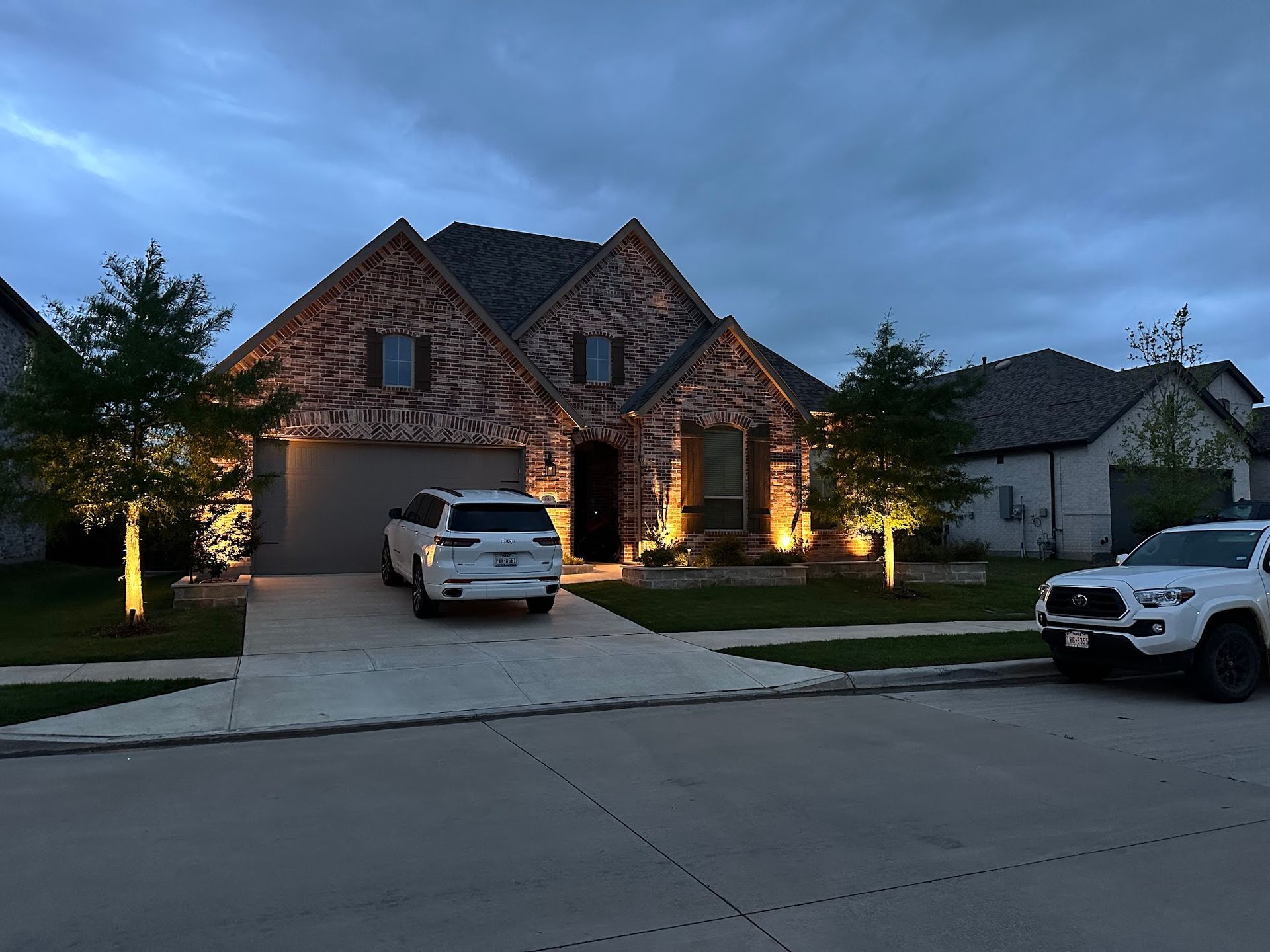 House at dusk with outdoor lighting, two vehicles in driveway.