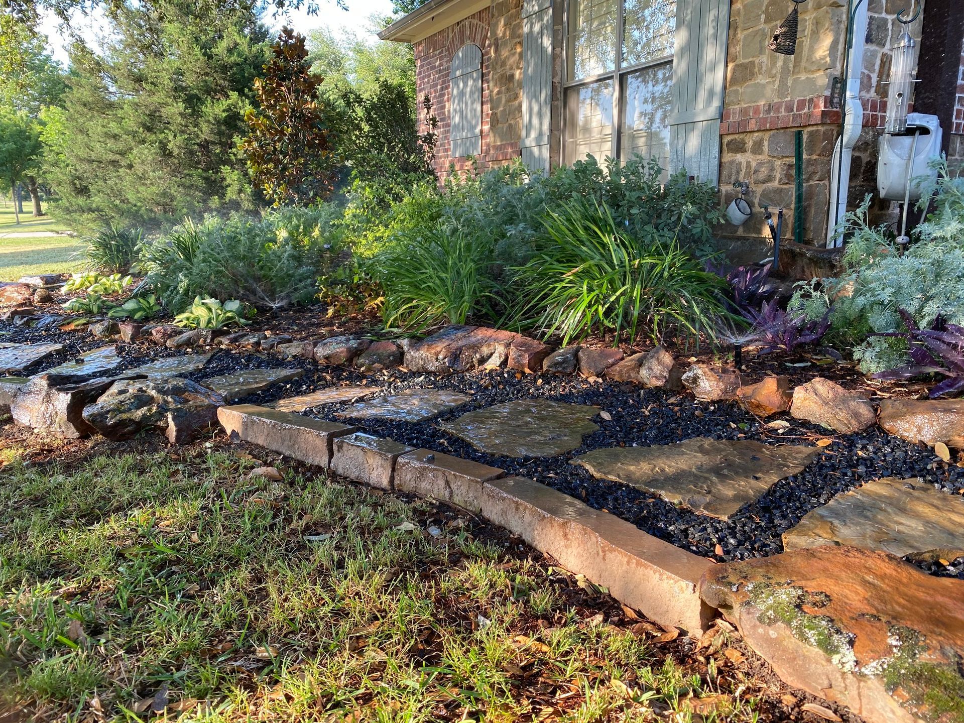 Stone pathway bordered by wood and rocks, leading to a brick house with overgrown garden beds.