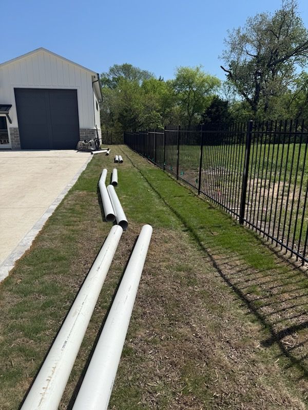 White PVC pipes on grass next to a building and black fence. Sunny day.