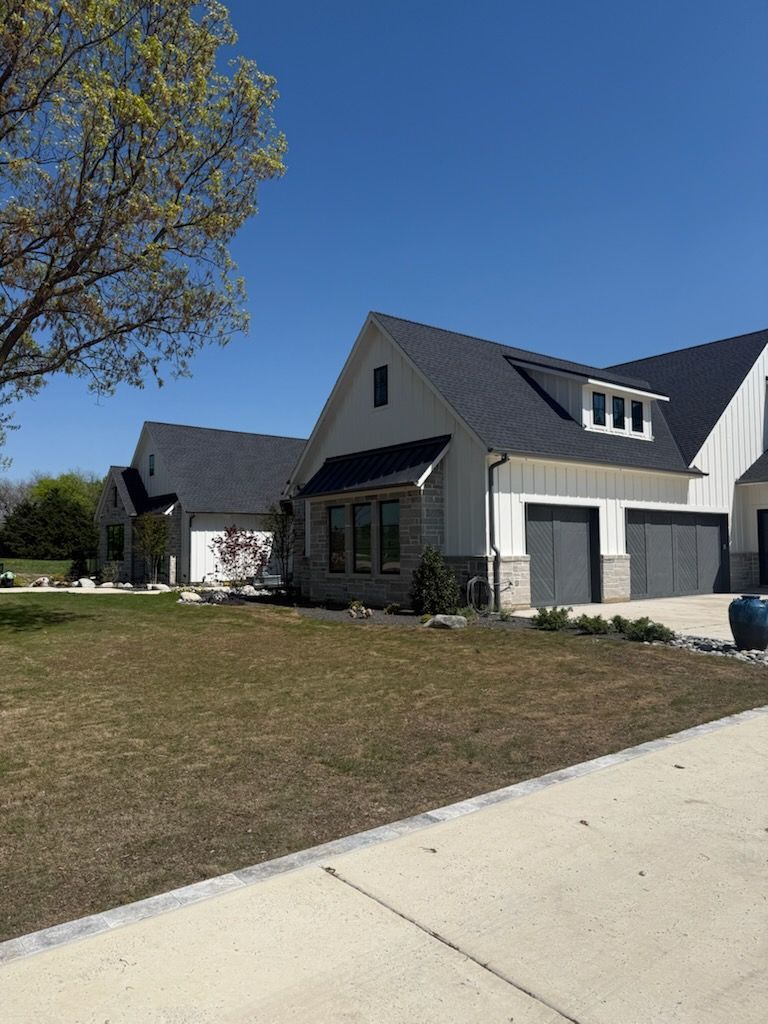Modern two-story house with gray roof and garage doors, white siding, stone accents, and a blue sky.