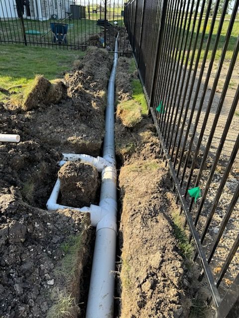 Trench with installed PVC piping along a black fence in a grassy yard.