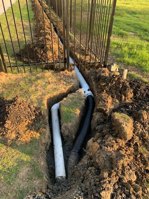 Trenches with white and black pipes under a metal fence; drainage system installation.