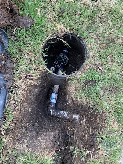 Open irrigation valve box and exposed PVC pipe in a grassy yard, dirt surrounding the hole.