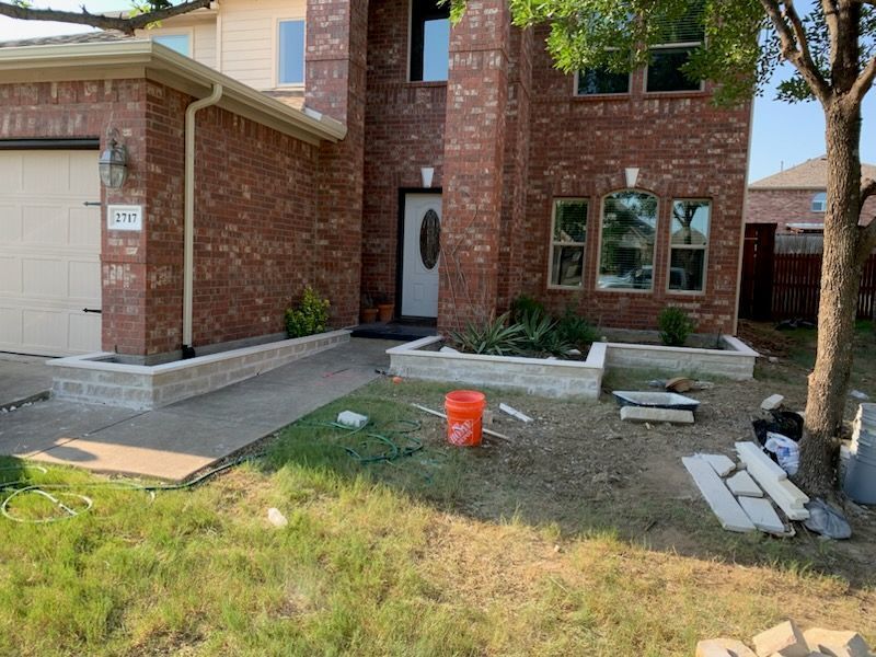 House exterior with brick facade, concrete walkway, and landscaping beds; construction materials and bucket present.