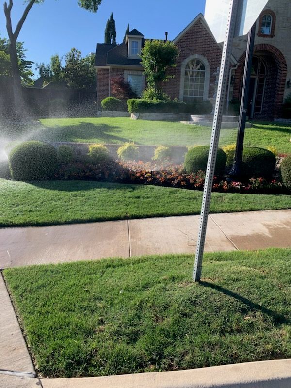 Lawn sprinkler watering a green lawn and bushes in front of a brick house on a sunny day.