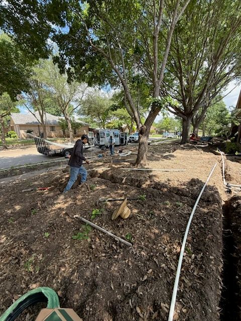 Construction site with workers digging trenches near trees and a road, on a sunny day.