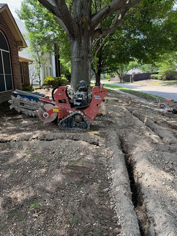 Trenching machine near a large tree, creating trenches in the dirt along a street.
