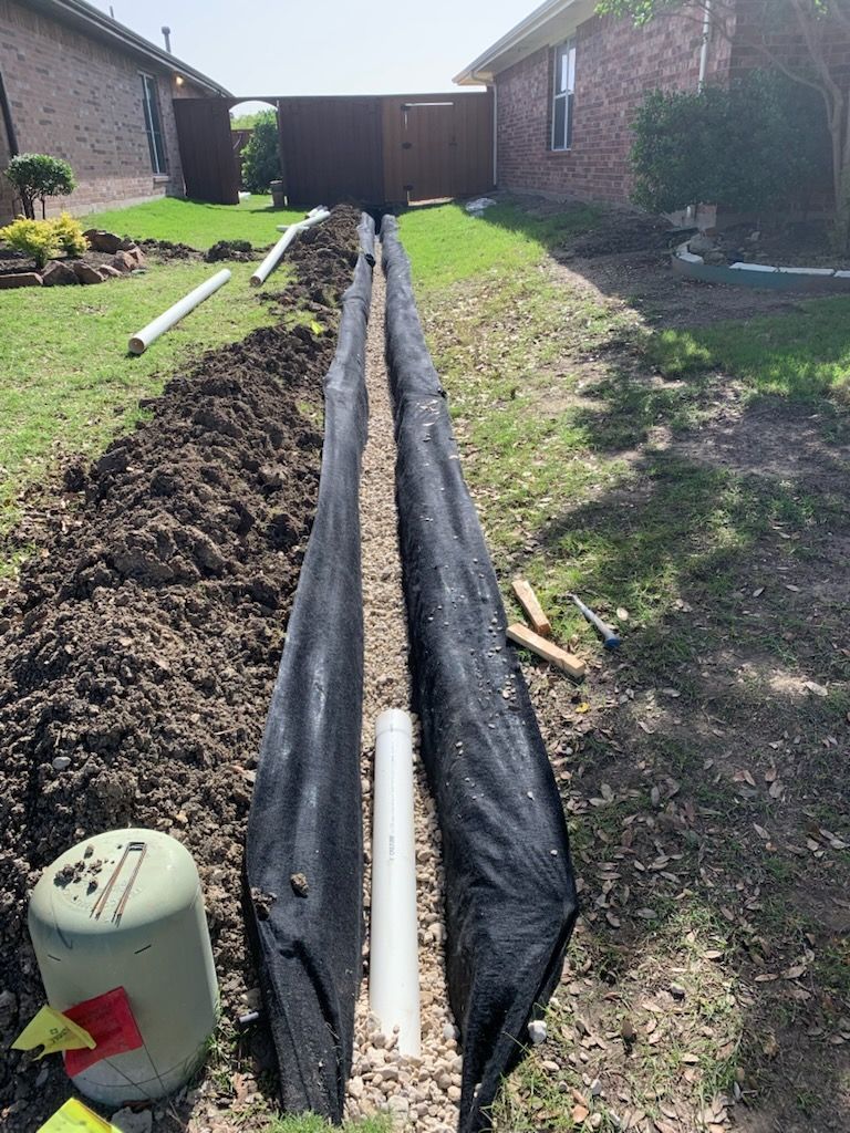 A drainage system being installed in a backyard. A trench filled with gravel and fabric contains white pipes.