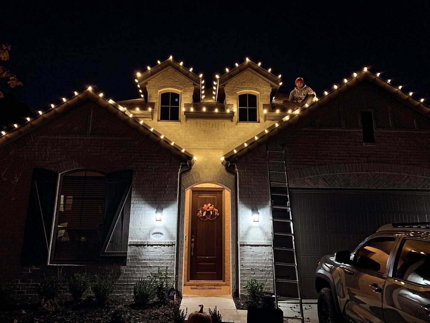 House decorated with string lights at night; person on roof, ladder nearby.