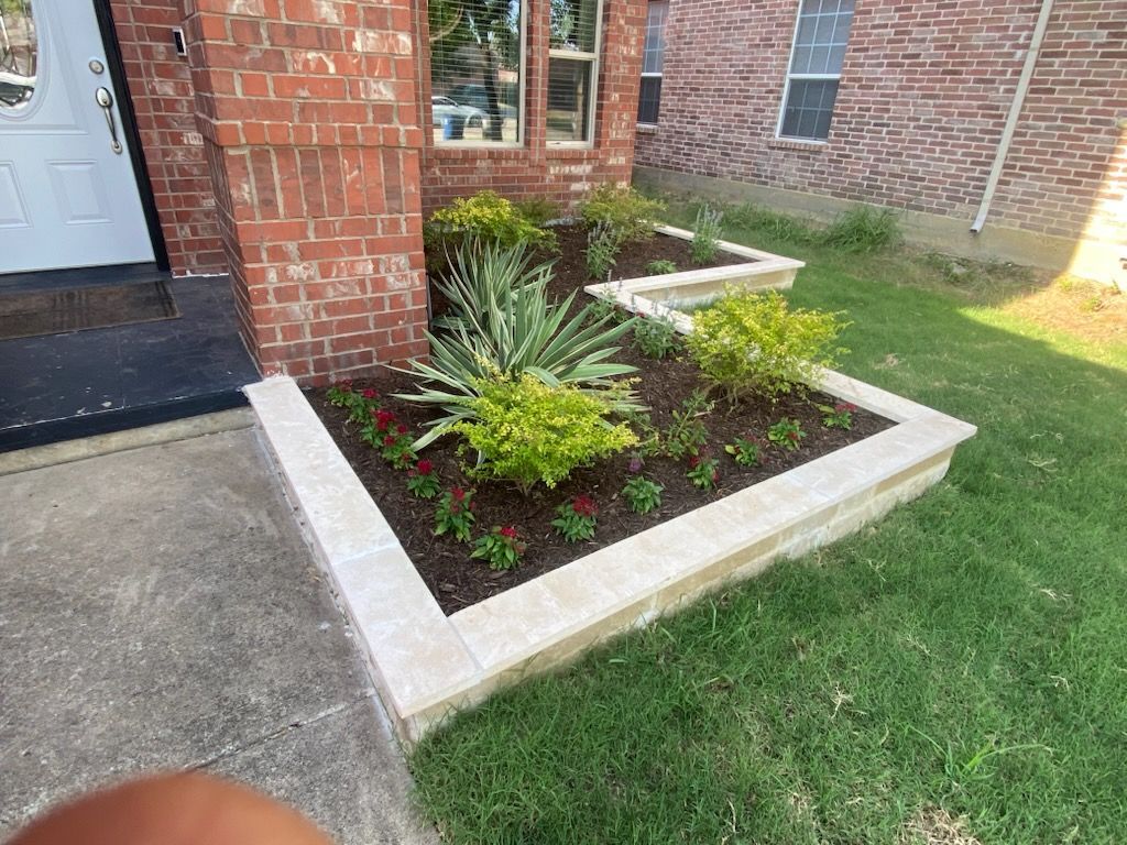 Stone-bordered garden bed with various plants, including a central agave, set against a brick pillar and green lawn.