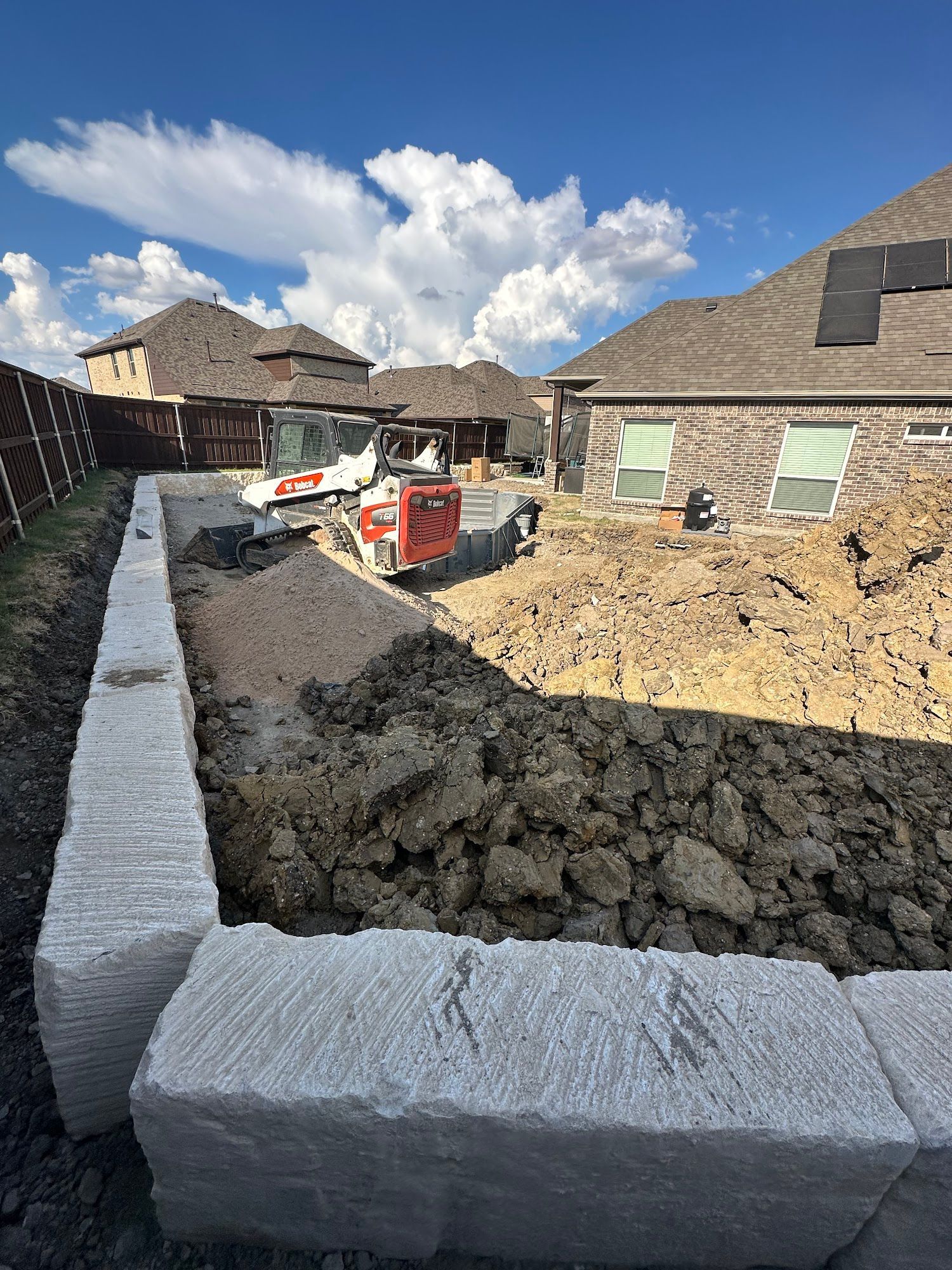 Construction site: Excavation of dirt inside a retaining wall; small excavator in the background; sunny day.