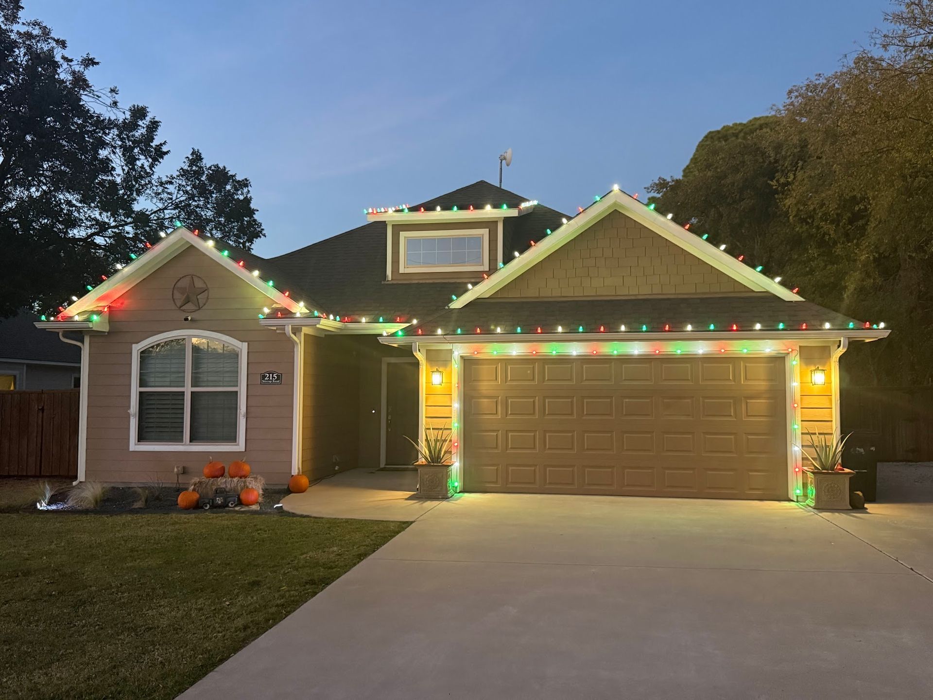 House with Christmas lights on roofline, front door, and garage door. Orange pumpkins decorate the yard.