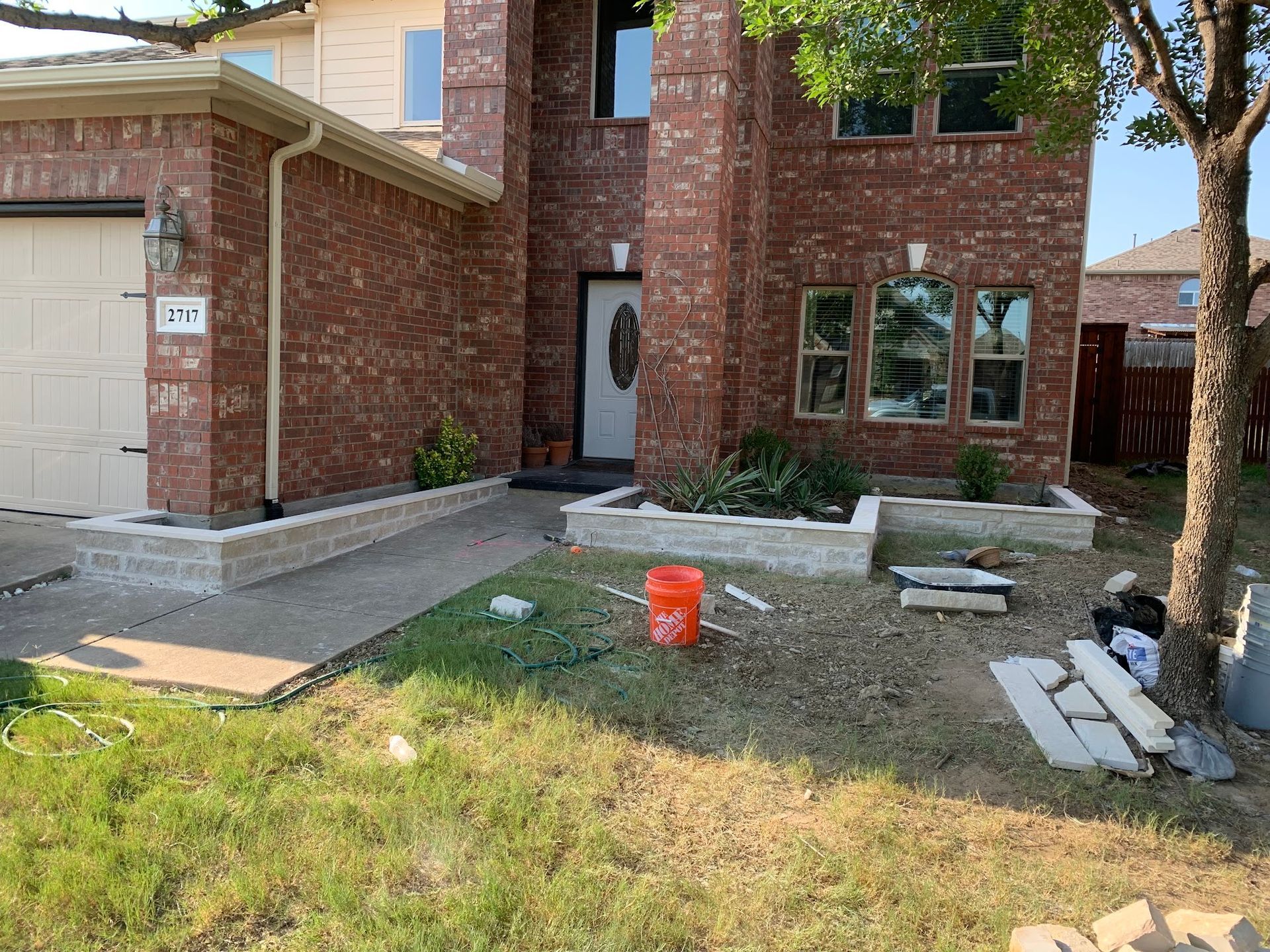 Brick house with front yard, concrete path, and flower beds. Construction materials and bucket visible.