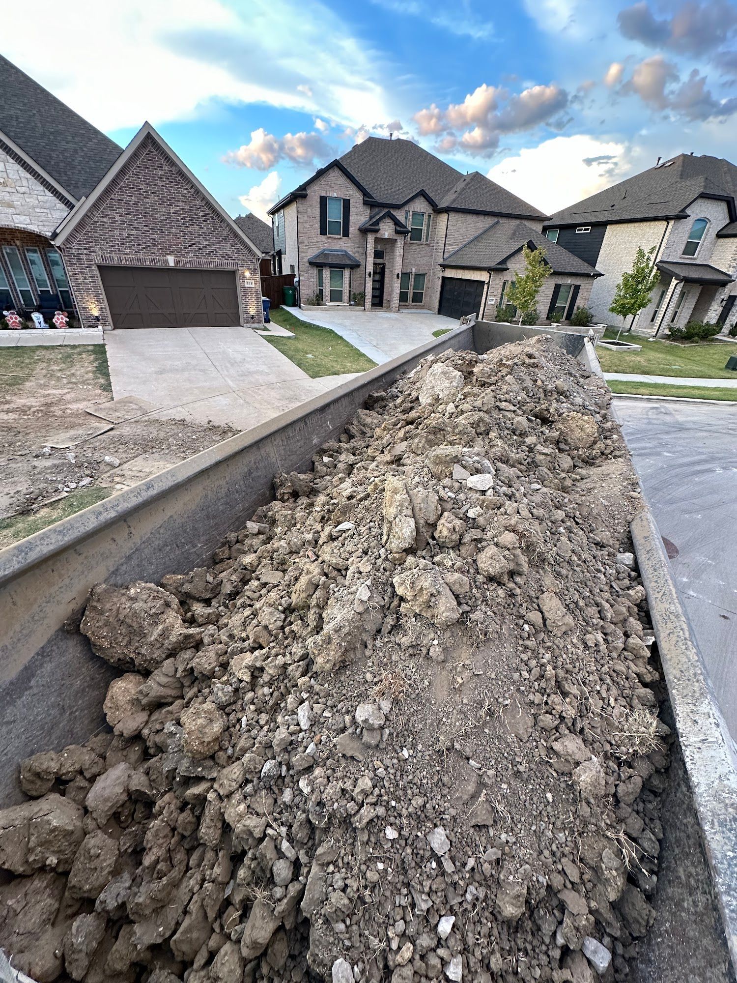 Dump truck bed filled with dirt on a residential street. Houses in background, blue sky.
