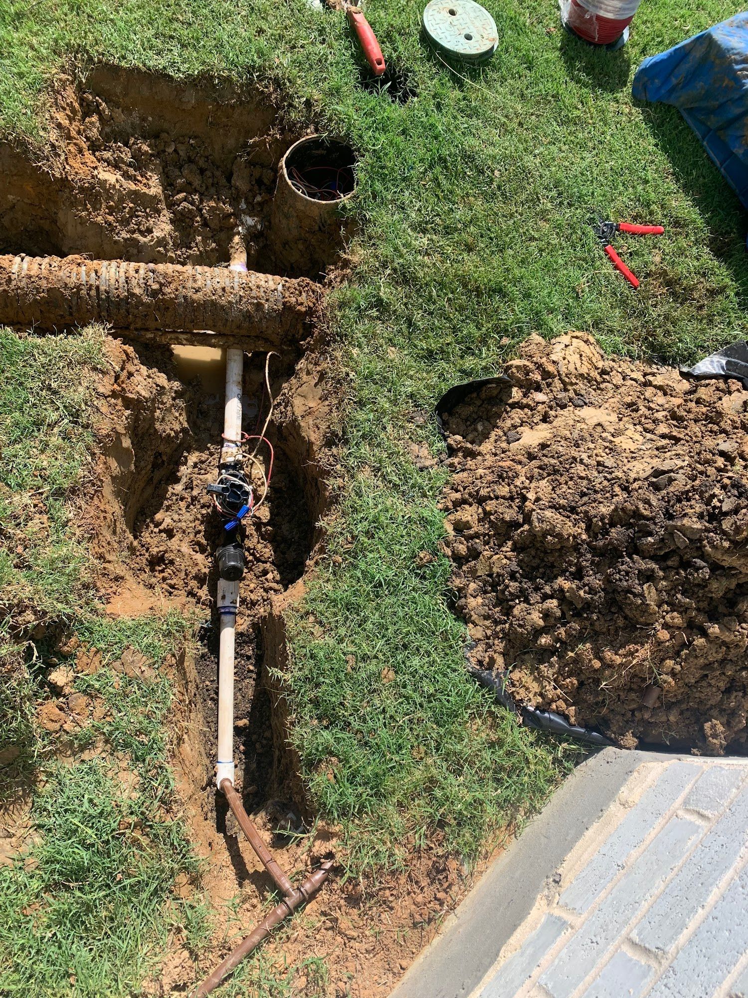 Irrigation system components exposed in a trench in a grassy yard, with tools nearby.