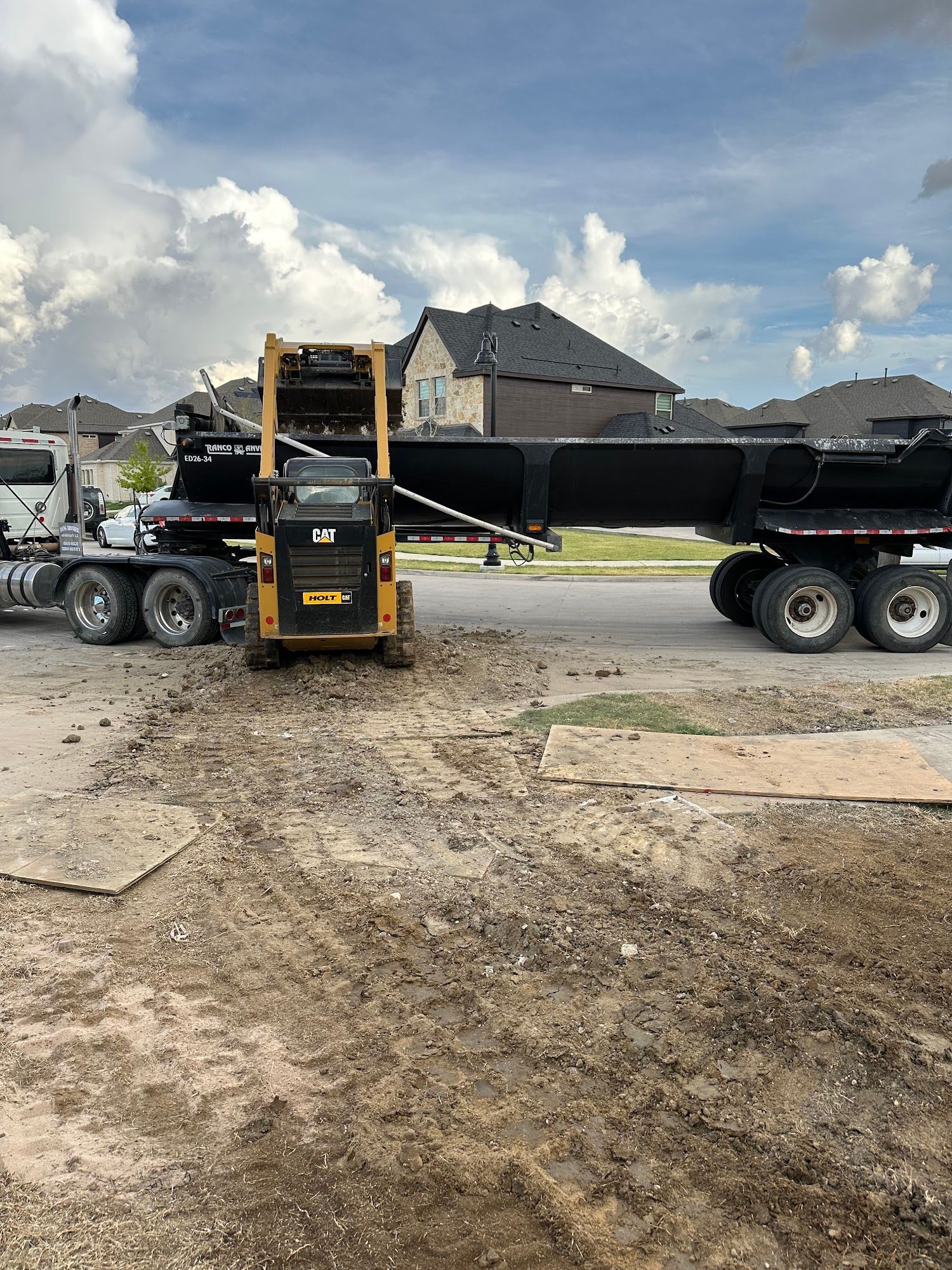 Yellow skid steer in front of a black trailer on a dirt construction site. Cloudy sky in the background.