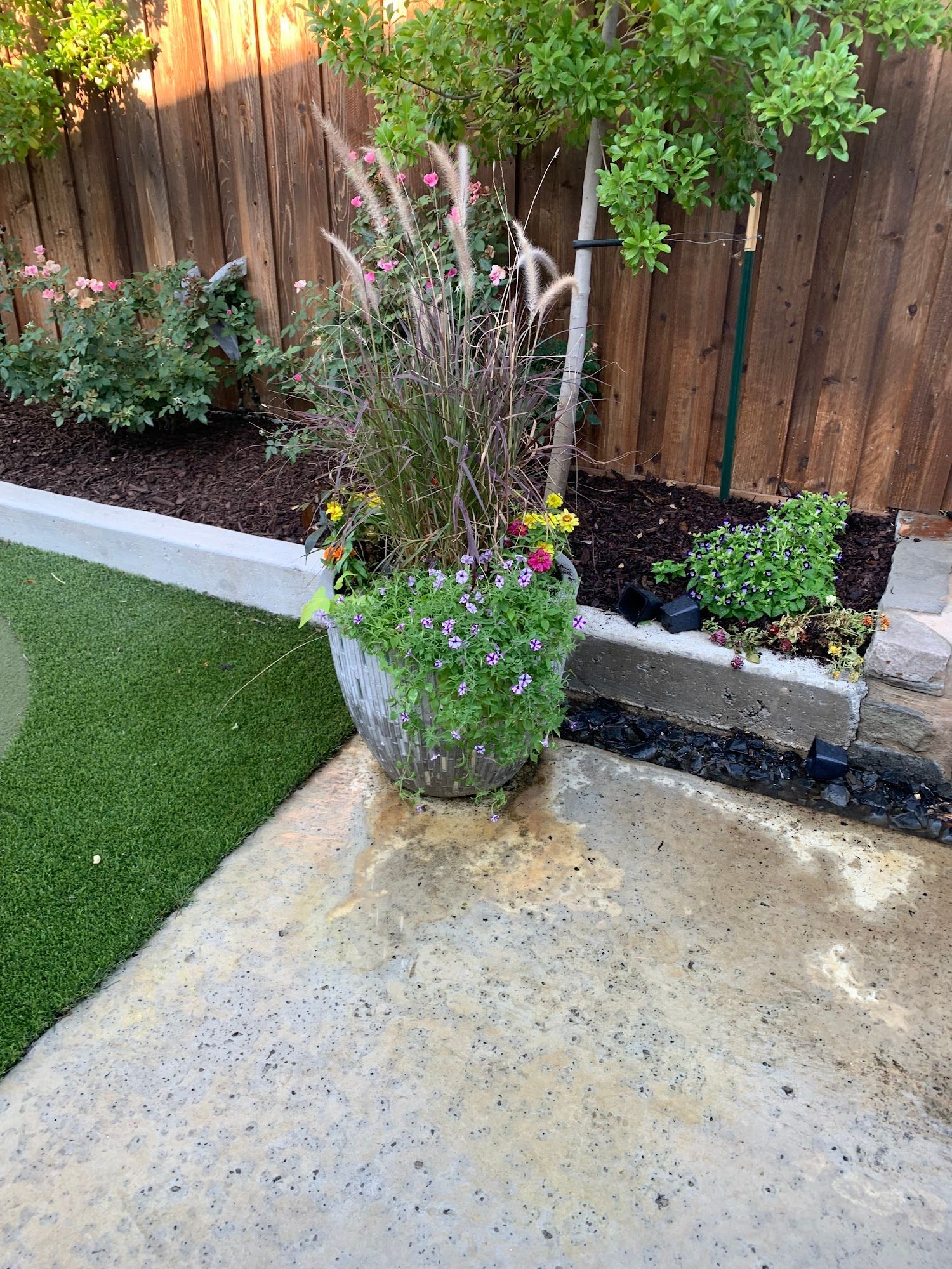 Stone planter with flowers, grasses, and small tree on a concrete patio next to artificial grass and a wooden fence.