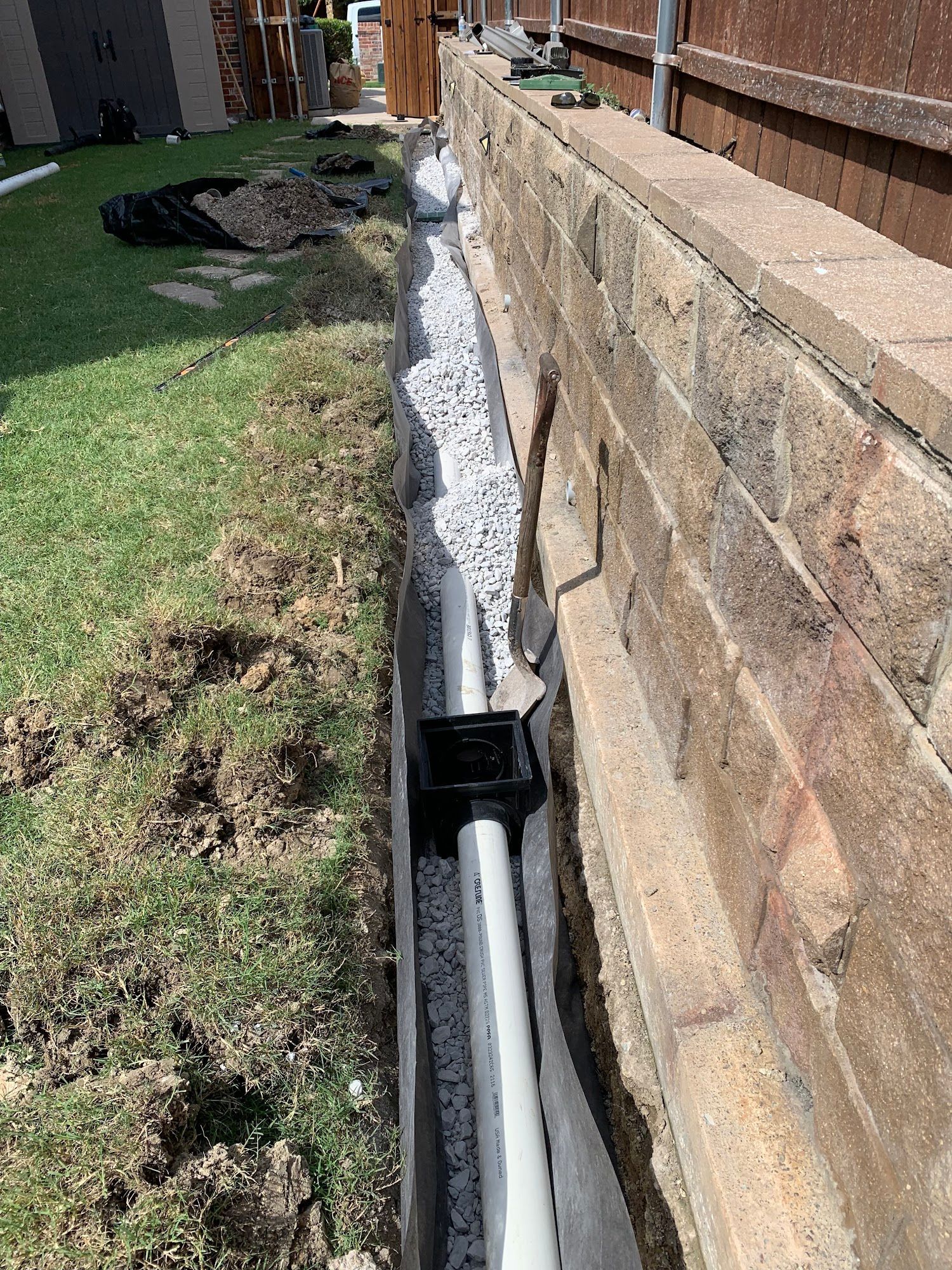 Trench drain installed against a brick wall, filled with gravel and connected to white piping, on a grassy lawn.