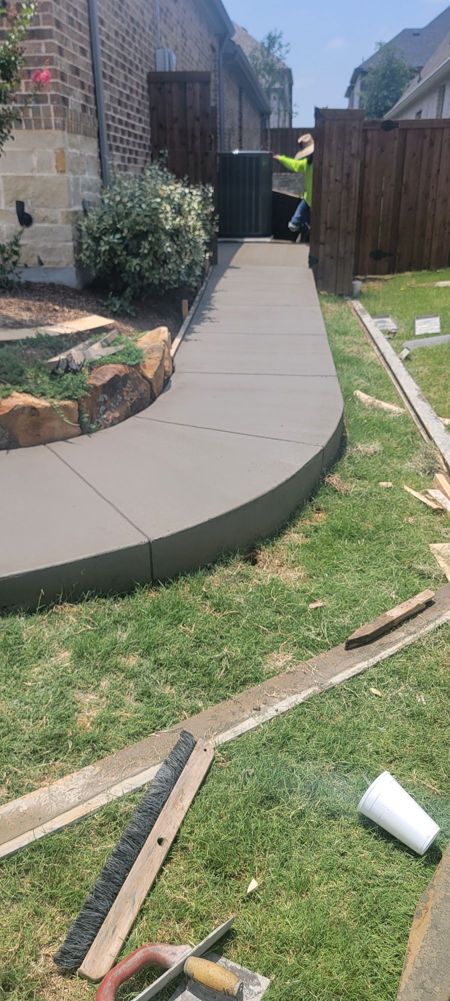 A newly poured concrete pathway curves through a green yard. Tools and forms are visible, indicating recent construction.