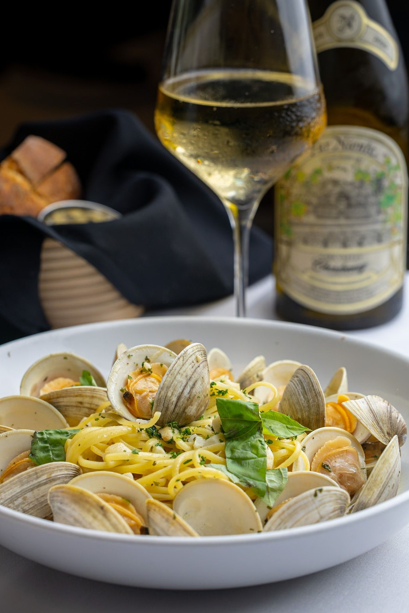 A bowl of clams and noodles with a glass of wine in the background.