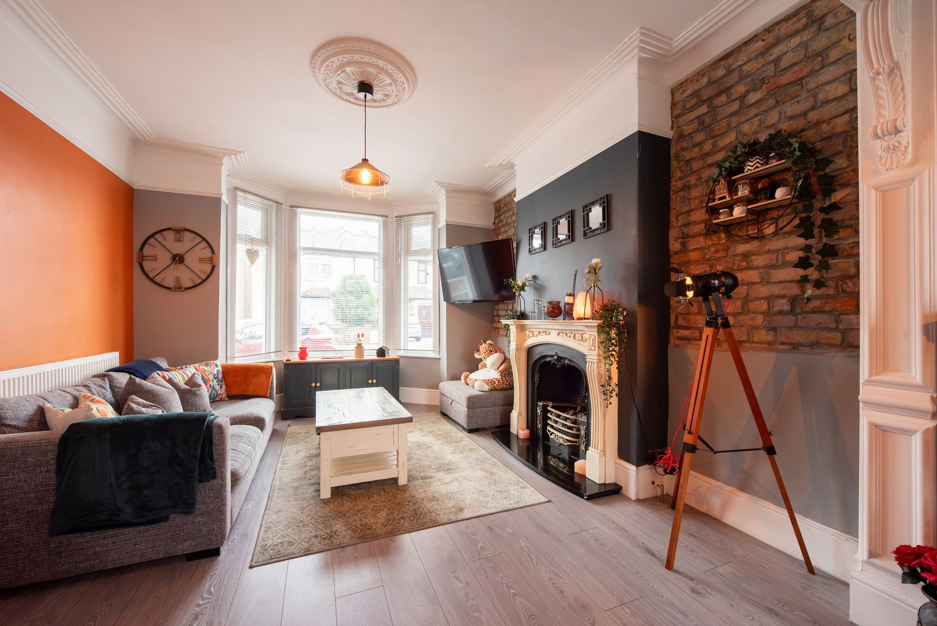 Living room with grey sofa, brick fireplace, orange accent wall, and tripod.