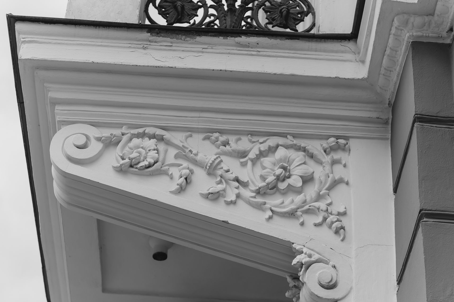 Black and white architectural detail of a building corner, showing ornate white plaster corbel with floral and scrollwork.