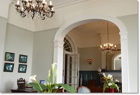 Elegant interior with archway, chandeliers, and muted colors. Features a table with flowers, and a glimpse into another room.