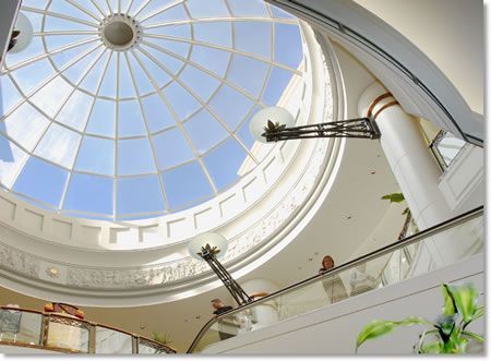 Inside view of a bright, modern building with a large glass dome skylight. People are visible on escalators and balconies.