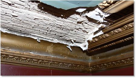 Damaged ceiling corner with peeling plaster, exposing wooden laths, gold decorative molding, and hints of red and turquoise paint.