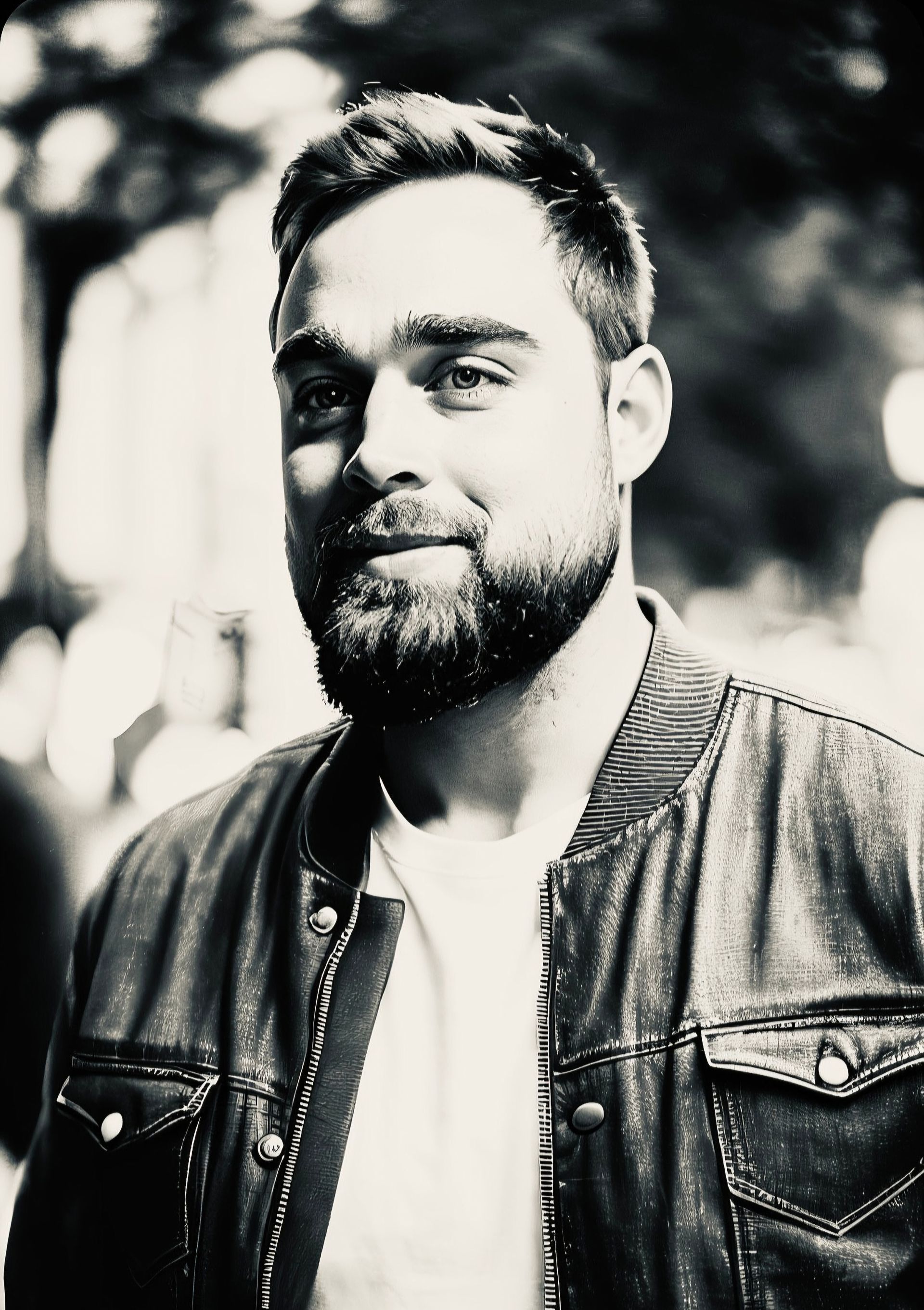 A man with a beard is wearing a leather jacket and a white shirt.