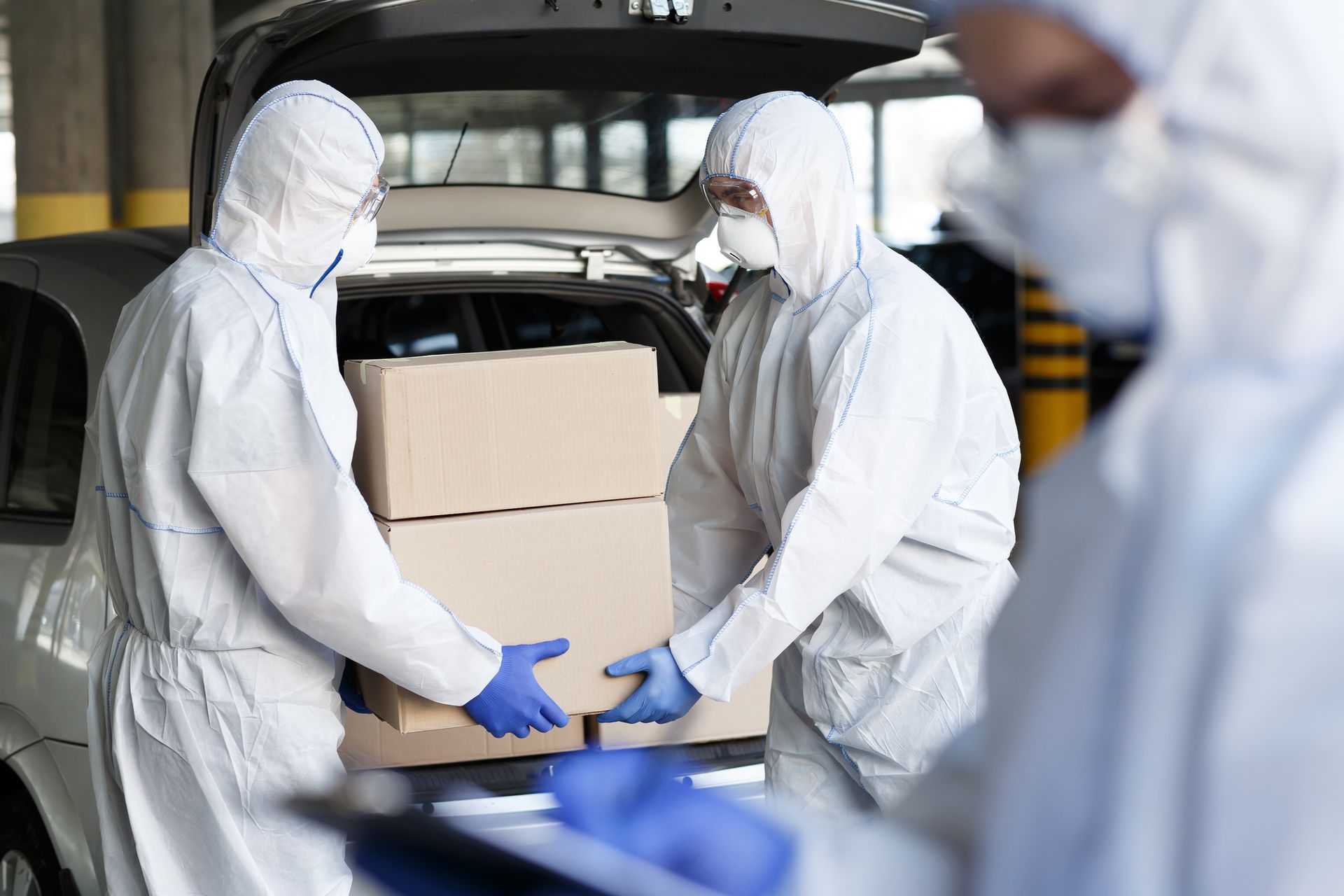 Two men in protective suits are loading boxes into the back of a car.