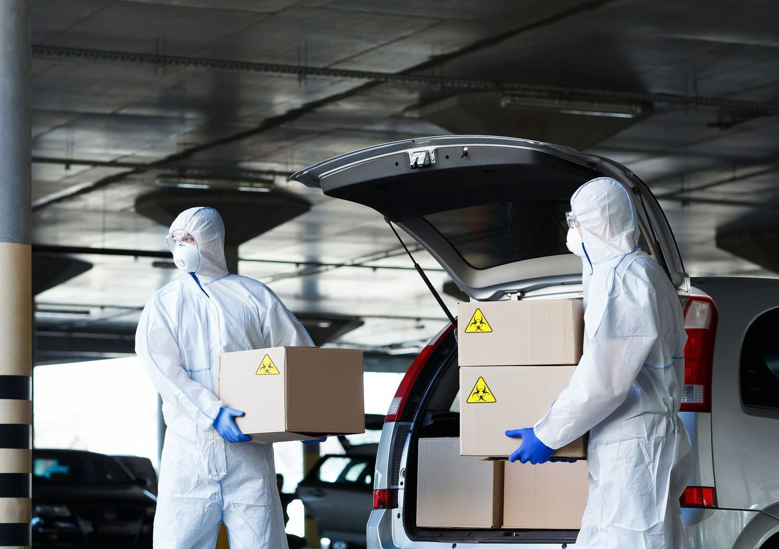 Two men in protective suits are loading boxes into the back of a car.