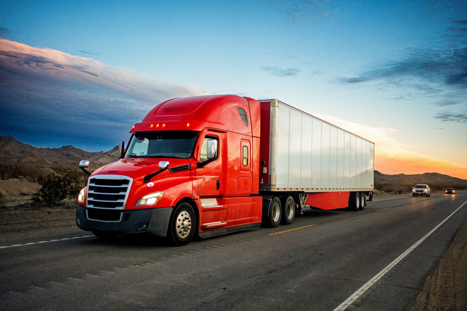 A red semi truck is driving down a highway at sunset.