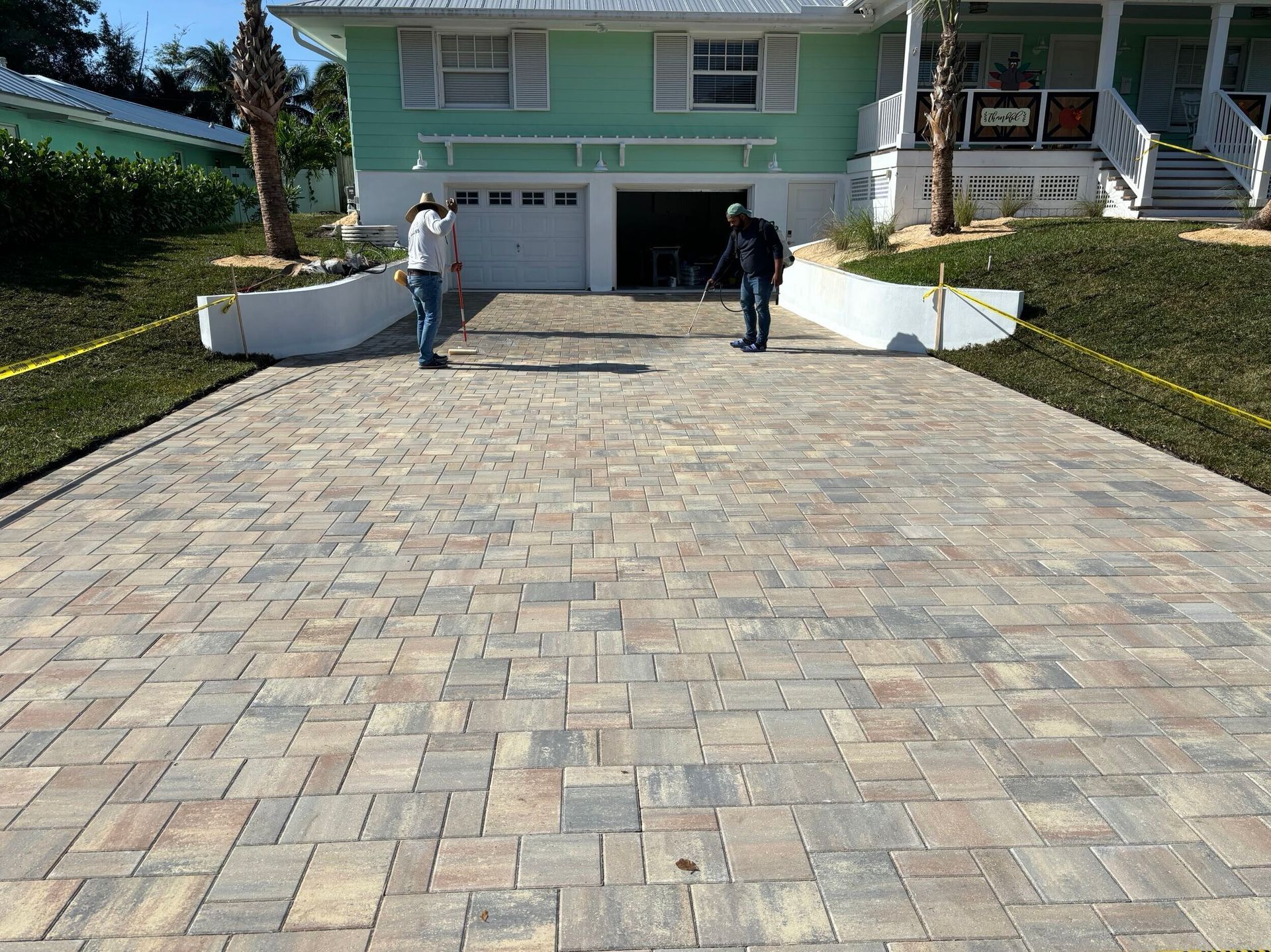 Driveway paved with multi-colored bricks in front of a light green house; two people are standing on the driveway.