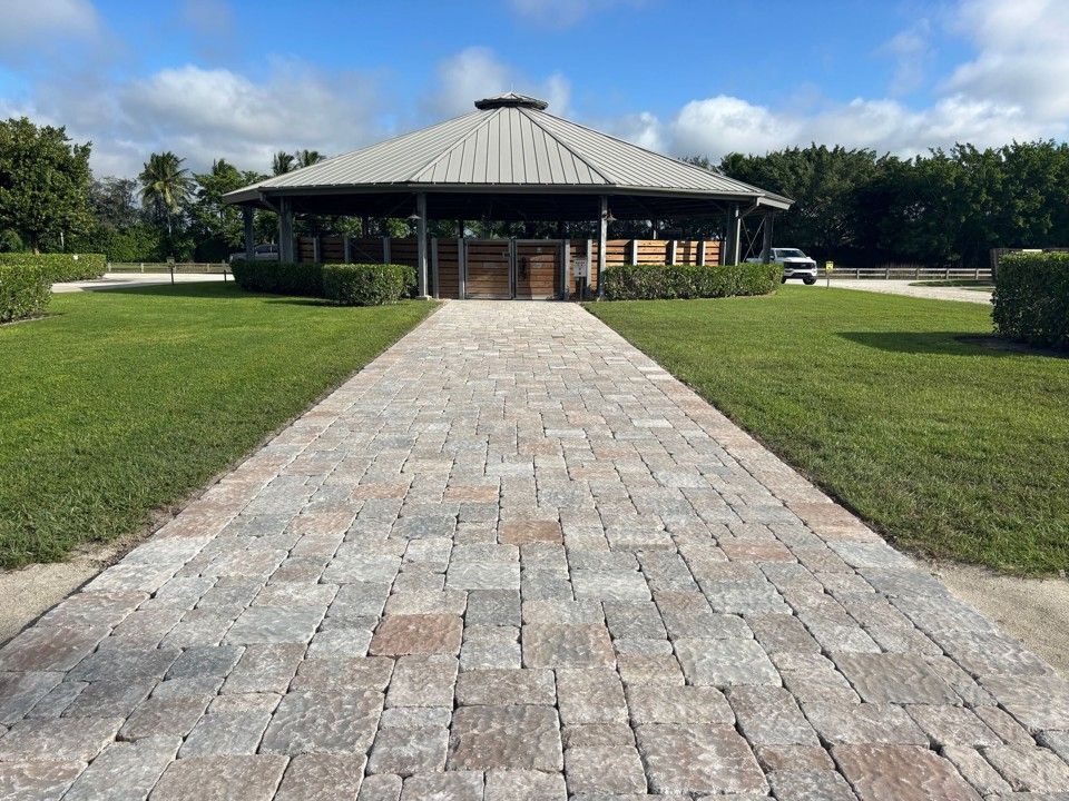 Brick path leads to open-air pavilion with metal roof, surrounded by green grass and shrubbery under a blue sky.