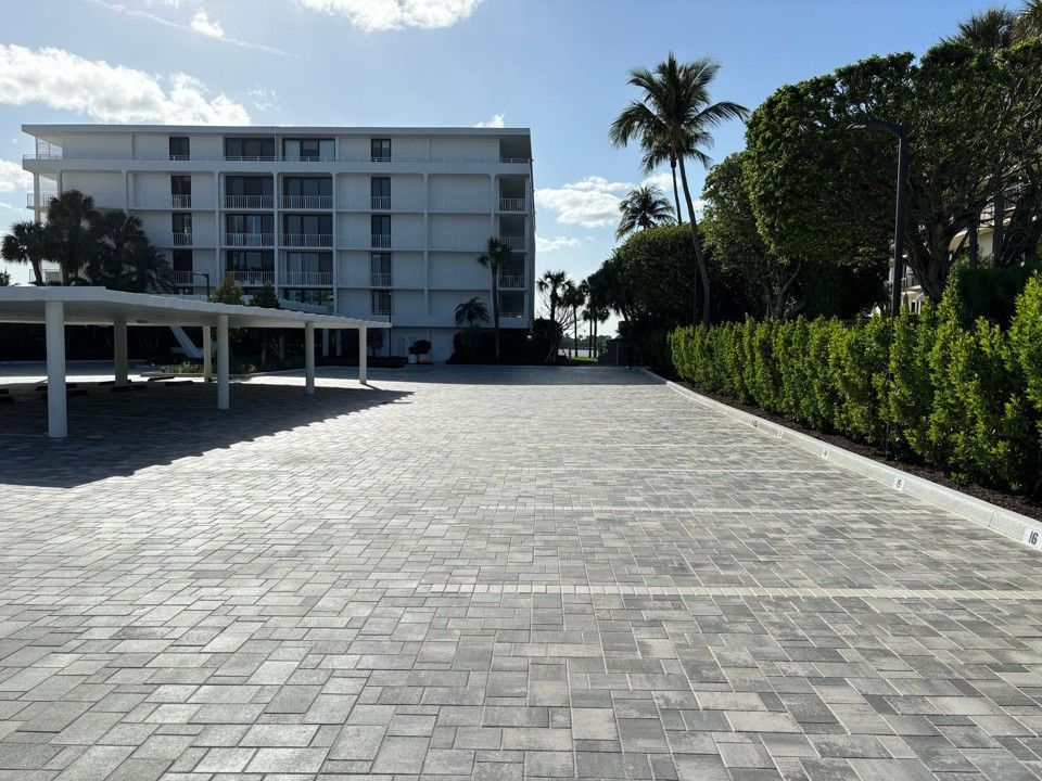 Brick paved parking lot with a white apartment building in the background. Green hedge and palm trees.
