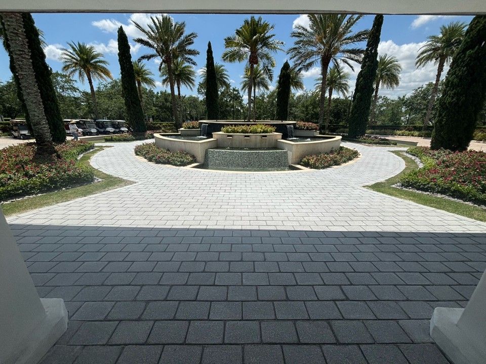 View from under a white structure leads to a fountain and palm trees in a sunny, paved courtyard.