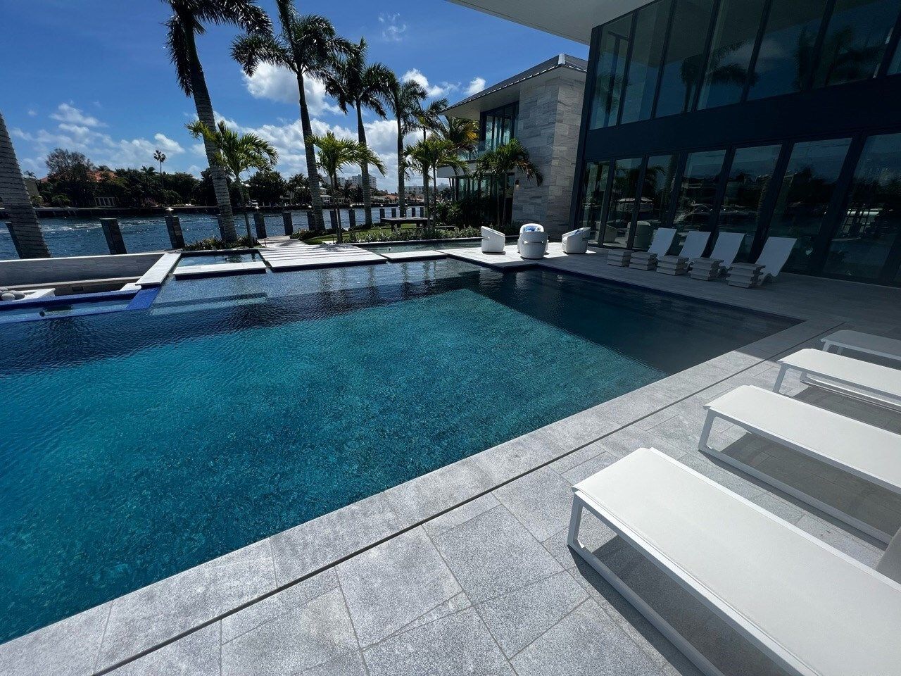 A modern pool surrounded by a patio and lounge chairs, with a mansion and palm trees in the background.