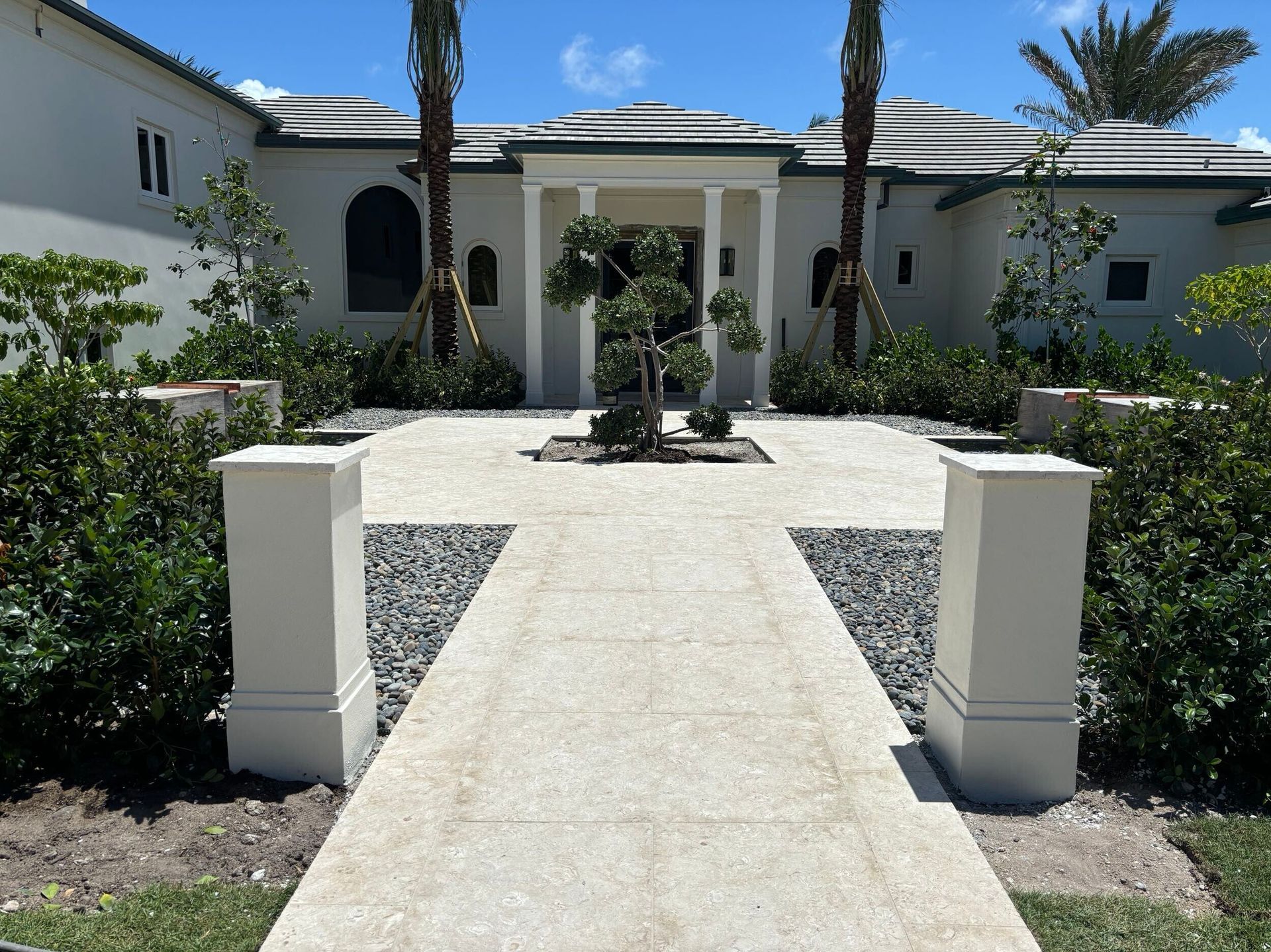 Stone path leads to a white house with columns, flanked by shrubs and palm trees, under a blue sky.