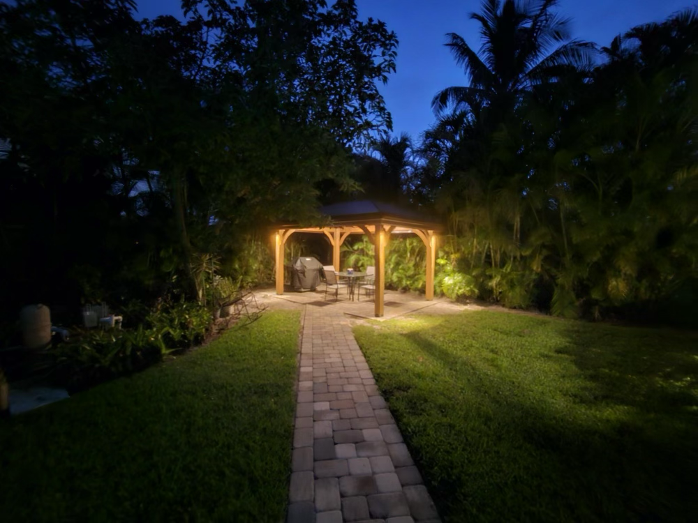 Nighttime view of a lighted wooden gazebo with a brick pathway and surrounding green lawn and foliage.